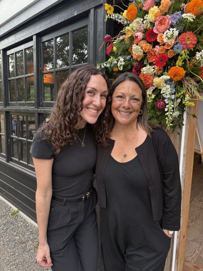 Photo of Diana and Jess at the wedding, smiling with a backdrop of colorful floral arrangements and tent lighting