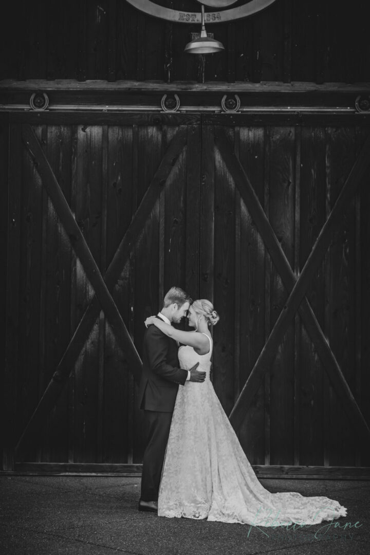 Jess and Tristan embrace in front of the rustic barn at The Kelley Farm in Bonney Lake, Washington, in a romantic black and white wedding portrait.